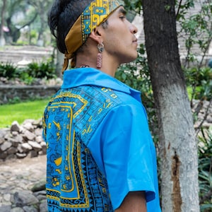 May include: A blue short-sleeved shirt with a yellow and black geometric pattern. The shirt has a collar and a button-up front. The person is wearing a yellow and black patterned headband.
