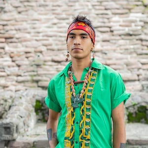 May include: A man wearing a green shirt with a yellow and black patterned design, a red and black bandana, and a beaded necklace with a green bird pendant. He is standing in front of a stone wall.