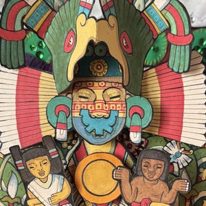 May include: A vibrant paper artwork featuring an Aztec figure with a detailed headdress and facial markings. Two figures flank the central character, holding a golden disc. The artwork uses bright greens, reds, yellows, and blues.