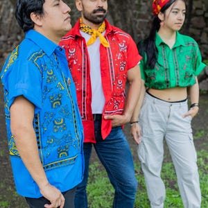 May include: Three people wearing colorful patterned shirts stand in front of a stone wall. The person on the left is wearing a blue shirt with yellow and green patterns. The person in the middle is wearing a red shirt with yellow and white patterns. The person on the right is wearing a green shirt with white and yellow patterns.