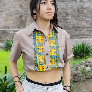 May include: A woman wearing a brown short-sleeved shirt with a yellow, blue, and black geometric pattern. The shirt is cropped and shows her midriff. She is also wearing white pants and has a bracelet on her wrist.