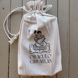 May include: A beige drawstring pouch made of fabric, featuring a black printed graphic of a seated figure on a platform and the text OR&Aacute;CULO CIHUATL&Aacute;N. The bag has a gathered top with two white rope cords extending from the sides for closure.