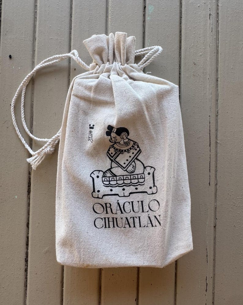 May include: A beige drawstring pouch made of fabric, featuring a black printed graphic of a seated figure on a platform and the text OR&Aacute;CULO CIHUATL&Aacute;N. The bag has a gathered top with two white rope cords extending from the sides for closure.