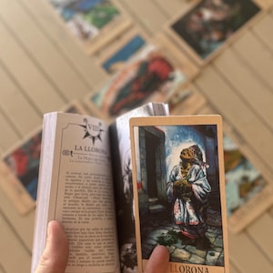 May include: A small book open to a page titled VIII LA LLORONA with Spanish text, held alongside a rectangular tarot card featuring a colourful illustration of a figure in a white robe looking upward, with the word LLORONA printed at the bottom.