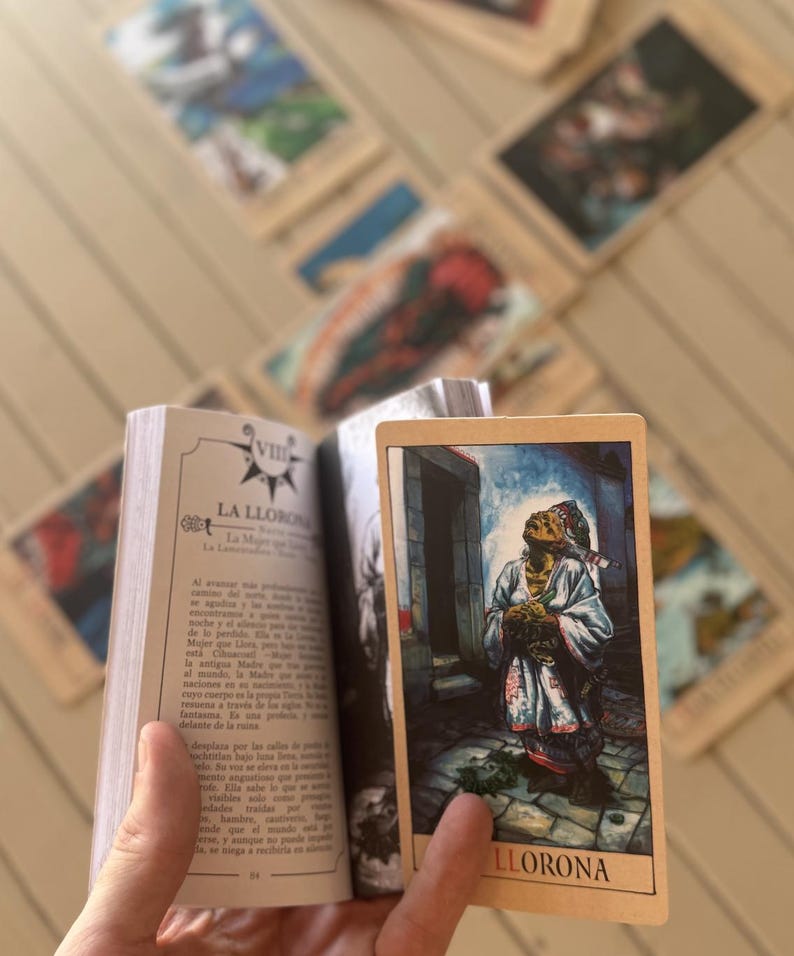 May include: A small book open to a page titled VIII LA LLORONA with Spanish text, held alongside a rectangular tarot card featuring a colourful illustration of a figure in a white robe looking upward, with the word LLORONA printed at the bottom.