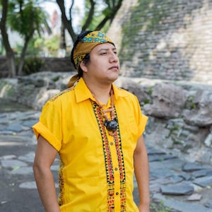 May include: A man wearing a yellow shirt with black and red geometric patterns and a brown headband with a blue and green pattern. He is wearing a necklace with a dark brown pendant.