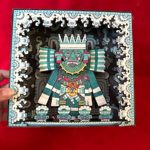 Könnte beinhalten: Eine 3D-Pop-up-Karte mit einer farbenfrohen, detaillierten Illustration einer Gottheit mit einer Krone, einem Federkopfschmuck und komplizierten Mustern. Die Gottheit ist von kleineren Figuren und einem dekorativen Rand umgeben.
