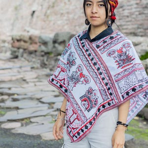 May include: A woman wearing a white poncho with red and black geometric patterns and a red headband. The poncho has a black collar and features a design of stylized figures. She is standing in front of a stone wall.