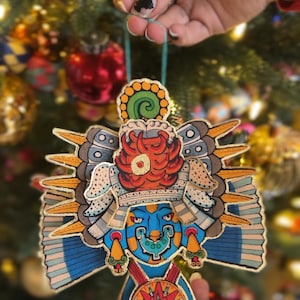 Puede incluir: Un adorno de papel colorido que representa una cara estilizada con una corona y alas. El adorno está decorado con detalles dorados y presenta un esquema de color azul, rojo, verde y amarillo.