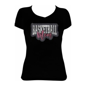 May include: Black V-neck t-shirt with the text "BASKETBALL Mom" in a rhinestone design. The word "BASKETBALL" is in silver rhinestones, and "Mom" is in pink rhinestones. The shirt is a solid black color.