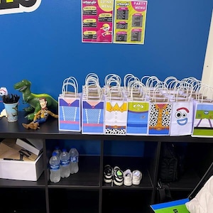 Puede incluir: Bolsas de regalo tem&aacute;ticas con dise&ntilde;os de personajes, dispuestas sobre una estanter&iacute;a negra. Las bolsas tienen tem&aacute;tica, con una pared azul de fondo. Tambi&eacute;n se ven personajes de Toy Story y men&uacute;s.