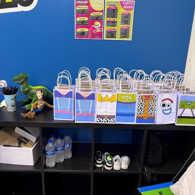 Puede incluir: Bolsas de regalo tem&aacute;ticas con dise&ntilde;os de personajes, dispuestas sobre una estanter&iacute;a negra. Las bolsas tienen tem&aacute;tica, con una pared azul de fondo. Tambi&eacute;n se ven personajes de Toy Story y men&uacute;s.