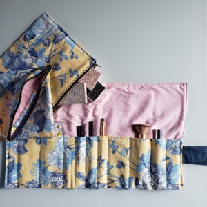 May include: A yellow and blue floral makeup roll-up with a pink lining. The roll-up has multiple compartments for storing makeup brushes and other makeup items.
