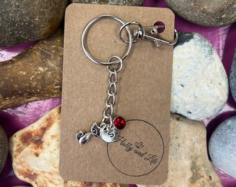 May include: A silver keychain with a charm of a cat and a red crystal. The keychain is attached to a brown card with the text "Holly and Lil'" printed on it.