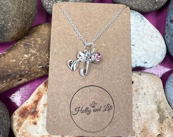 May include: A silver necklace with a charm of the letter J and a pink crystal. The necklace is displayed on a brown card with the text "Holly and Lili" in a circle.