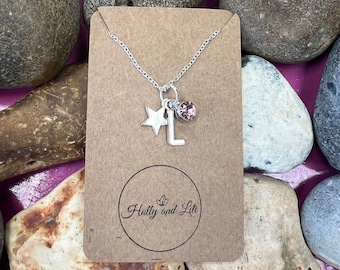 May include: A silver necklace with a star charm, a letter L charm, and a pink crystal bead. The necklace is displayed on a brown card with the text "Holly and Lili" in a circle.