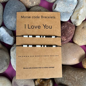 Puede incluir: Dos pulseras de cuentas blancas y negras con un mensaje en código Morse. El mensaje es "Te amo". Las pulseras están en una tarjeta marrón con el texto "Pulseras de código Morse" y "Pulsera de código Morse con un mensaje oculto".