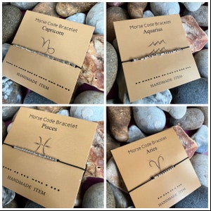 Puede incluir: Cuatro tarjetas marrones con pulseras de cuerda negra y cuentas plateadas. Cada tarjeta presenta un signo del zodíaco diferente: Capricornio, Acuario, Piscis y Aries. El texto en cada tarjeta dice "Pulsera de código Morse" seguido del signo del zodíaco. La parte inferior de cada tarjeta dice "ARTESANÍA".