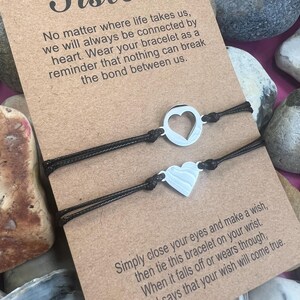 Peut inclure: Deux bracelets assortis avec des cordons noirs et des breloques en forme de cœur argenté, présentés sur une carte marron avec le mot "Sisters" et un message sur le lien. La carte est entourée de pierres.