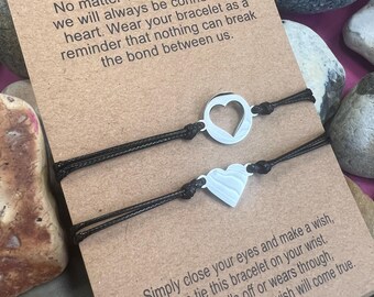 Può includere: Due braccialetti abbinati con cordini neri e ciondoli a forma di cuore argentato, presentati su una carta marrone con la parola "Sisters" e un messaggio sul legame. La carta è circondata da pietre.