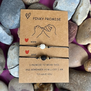 Puede incluir: Dos pulseras de cuerda negra con piedras de lava y cuentas blancas. Las pulseras están atadas juntas con una tarjeta que dice "Pinky Promise" y tiene un dibujo de dos manos haciendo una promesa meñique. La tarjeta también dice "mine" y "your" y "Prometo llevar esto y recordar lo afortunado que soy de tenerte."
