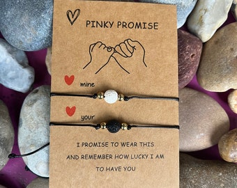 Puede incluir: Dos pulseras de cuerda negra con piedras de lava y cuentas blancas. Las pulseras están atadas juntas con una tarjeta que dice "Pinky Promise" y tiene un dibujo de dos manos haciendo una promesa meñique. La tarjeta también dice "mine" y "your" y "Prometo llevar esto y recordar lo afortunado que soy de tenerte."