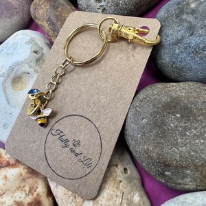 May include: Gold bee keyring with a blue gem on its back. The keyring is attached to a brown card with the text "Holly and Lil" in a circle.