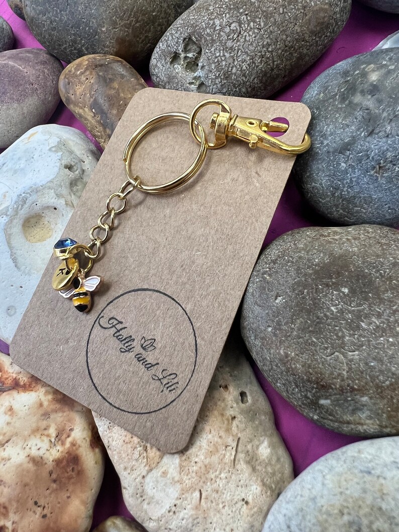 May include: Gold bee keyring with a blue gem on its back. The keyring is attached to a brown card with the text "Holly and Lil" in a circle.
