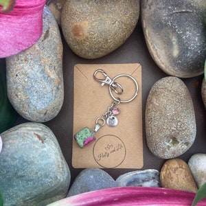 Pode incluir: Um chaveiro de prata com um charme de pedra verde e rosa, um charme inicial de prata e dois charmes rosa e prata. O chaveiro está em um cartão marrom com o texto "Holly and Lili" em branco.