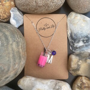 May include: A silver chain necklace with a pink bubble tea charm, a silver letter M charm, and a blue crystal charm. The necklace is on a brown card with the text "Holly and Lil" in white.