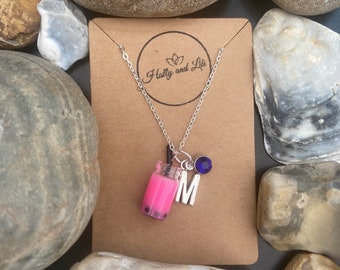 May include: A silver chain necklace with a pink bubble tea charm, a silver letter M charm, and a blue crystal charm. The necklace is on a brown card with the text "Holly and Lil" in white.