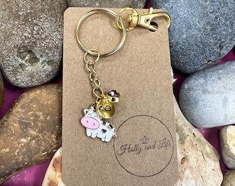 Könnte beinhalten: Goldener Schlüsselanhänger mit einem Kuh-Anhänger und einem rosafarbenen Strassstein. Der Schlüsselanhänger ist an einer braunen Karte mit dem Text "Holly and Lili" in einem Kreis befestigt.