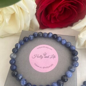 Könnte beinhalten: Ein blaues Edelstein-Armband in einer weißen und rosafarbenen Geschenkbox. Die Box hat einen Aufkleber mit dem Text "Holly and Lili" und der Website "hollyandlilii.etsy.com". Der Aufkleber enthält auch den Text "Folge uns: @hollyandlilii".