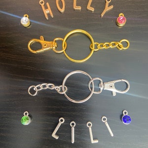 May include: Two keychains with gold and silver rings, chains, and clasps. The gold keychain has a pink rhinestone charm and the silver keychain has a green rhinestone charm. The letters "HOLLY" are spelled out in gold charms and the letters "LILI" are spelled out in silver charms. There are also blue and clear rhinestone charms.
