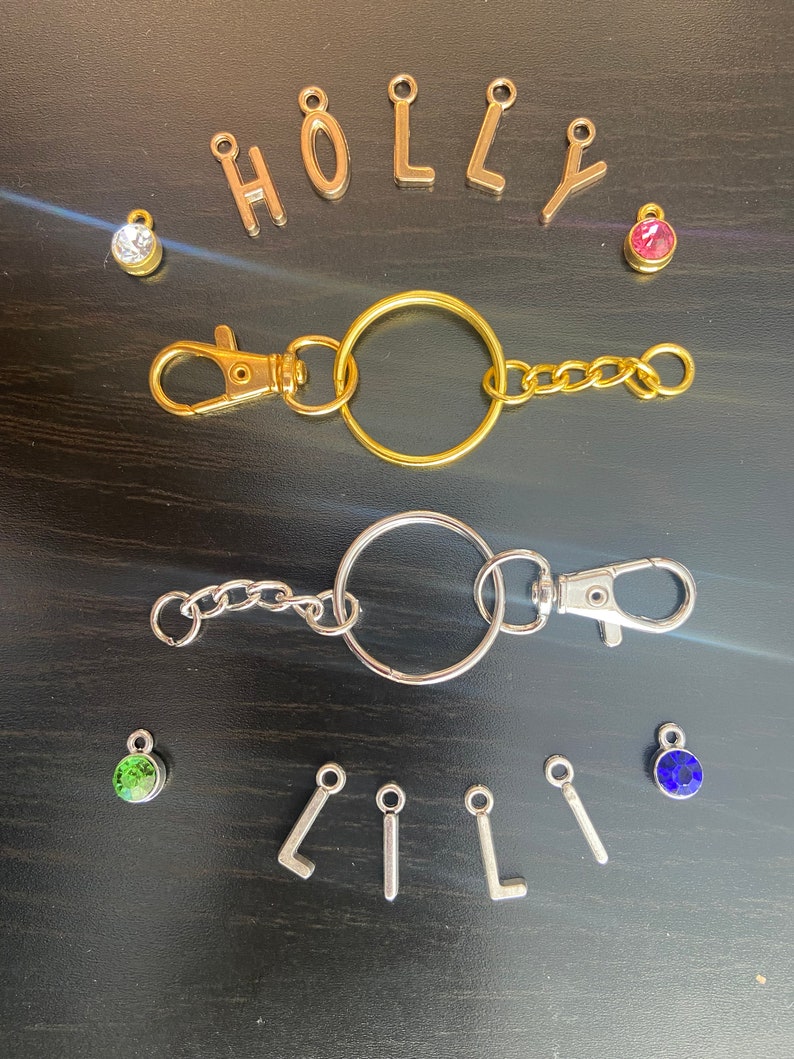May include: Two keychains with gold and silver rings, chains, and clasps. The gold keychain has a pink rhinestone charm and the silver keychain has a green rhinestone charm. The letters "HOLLY" are spelled out in gold charms and the letters "LILI" are spelled out in silver charms. There are also blue and clear rhinestone charms.