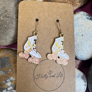 May include: A pair of gold-tone roller skate earrings. The earrings feature white roller skates with pink and yellow accents. The wheels are pink. The earrings are displayed on a brown card with the text "Hally and Lili".