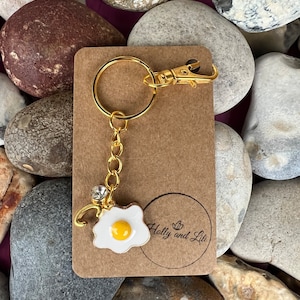 May include: Gold keyring with a fried egg charm and a clear rhinestone. The keyring is on a brown card with the text "Holly and Lili" printed on it.