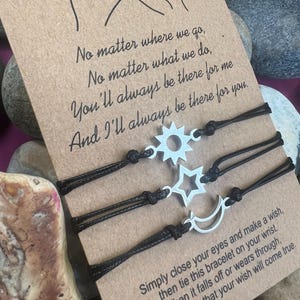 May include: Three wish bracelets with black cords and silver charms shaped like a sun, star, and crescent moon. The bracelets are displayed on a card with the text "No matter where we go..." and "Simply close your eyes and make a wish..."