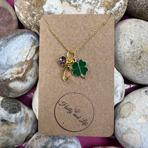 Può includere: Una collana con catena dorata con un ciondolo a forma di quadrifoglio verde e un ciondolo di cristallo rosa. La collana è esposta su una carta marrone con il testo "Holly and Lily" in un cerchio.