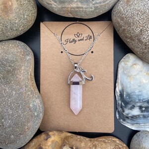 Puede incluir: Un collar de cadena plateada con un colgante de cristal rosa y un dije plateado en forma de la letra "J". El collar se muestra en una tarjeta marrón con el texto "Holly and Lili" impreso en ella.