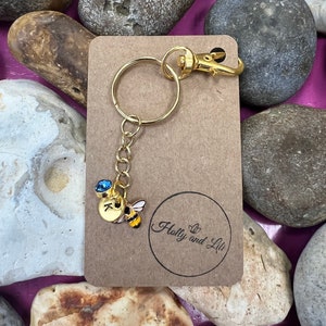 May include: A gold keyring with a small bee charm and a blue crystal. The charm has the letter "K" on it. The keyring is on a brown card with the text "Holly and Lili" in a circle.