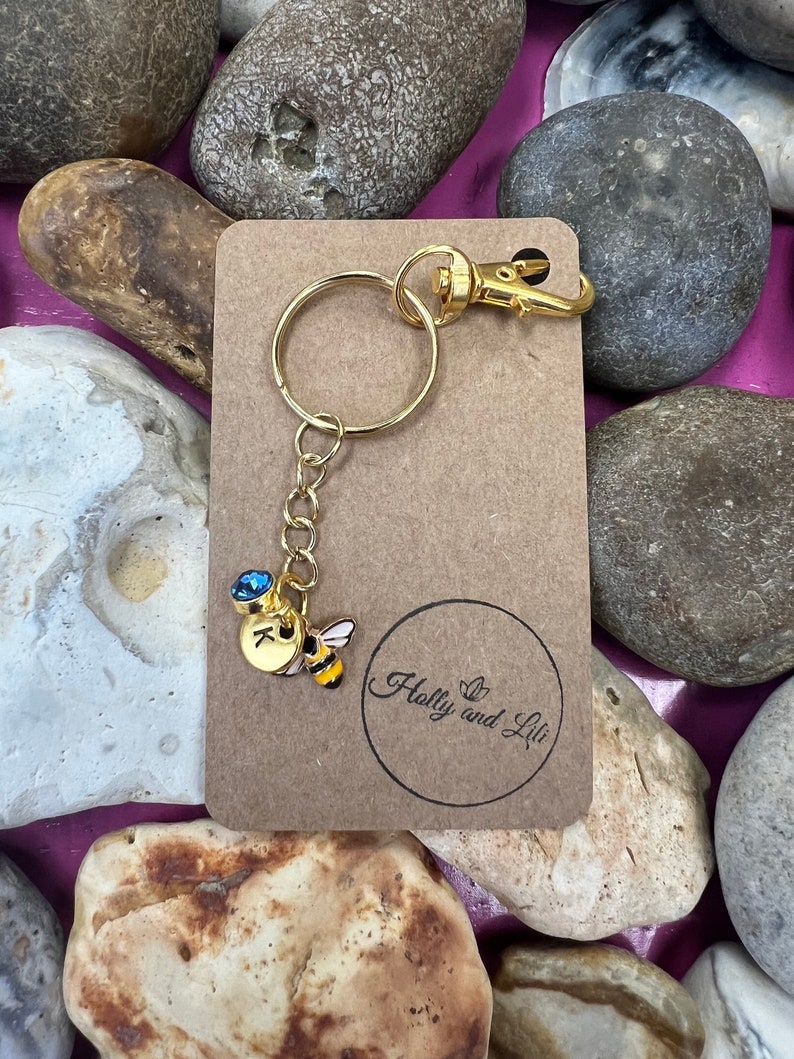 May include: A gold keyring with a small bee charm and a blue crystal. The charm has the letter "K" on it. The keyring is on a brown card with the text "Holly and Lili" in a circle.