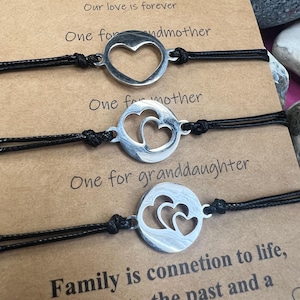 Puede incluir: Tres pulseras de deseos con cordones negros y dijes plateados con forma de corazón. El texto en la tarjeta dice "3 GENERACIONES" y "Nuestro amor es para siempre". Las pulseras se presentan en una tarjeta marrón con texto sobre la familia.
