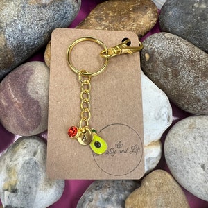 May include: A gold keychain with a charm of an avocado with a green pit and a red stone. The keychain also has a gold letter 'A' charm. The keychain is on a brown card with the text "Crafty and Lil" printed on it.