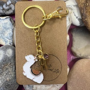 Op de afbeelding: Gouden sleutelhanger met een witte lama-hanger, een roze strasssteen en een gouden hanger met de letter "H". De sleutelhanger is bevestigd aan een bruine kaart met de tekst "Go and Live".