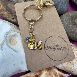 May include: Gold keyring with a blue rhinestone, a gold charm with the letter K, and a gold bee charm. The keyring is on a brown card with the text "Holly and Lili" printed on it.