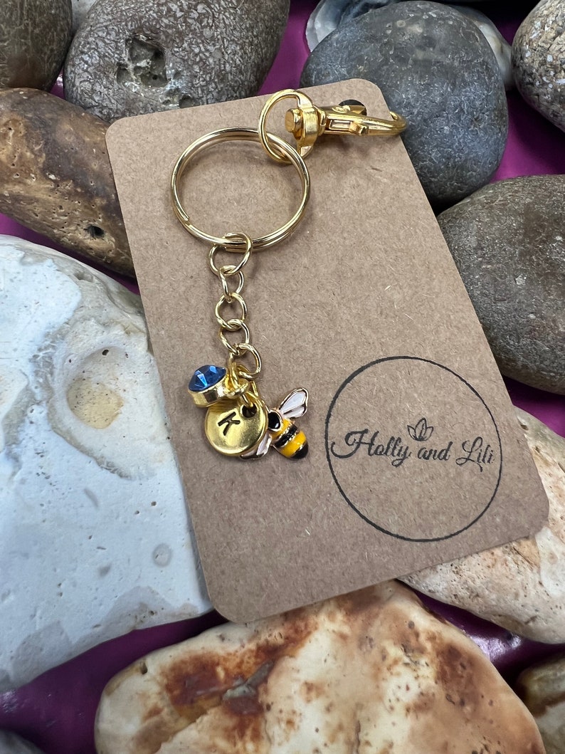 May include: Gold keyring with a blue rhinestone, a gold charm with the letter K, and a gold bee charm. The keyring is on a brown card with the text "Holly and Lili" printed on it.