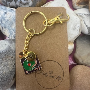 May include: Gold keychain with a green and red record player charm. The charm has a gold button and a small gold bead. The keychain also has a gold chain and a gold clasp. The keychain is on a brown card with the text "Holly and Lily" in a circle.