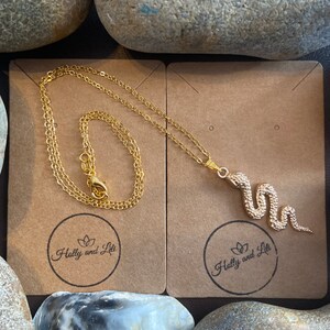 May include: A gold snake necklace with a delicate chain. The necklace is displayed on a brown card with the text "Holly and Liti" in a circle.
