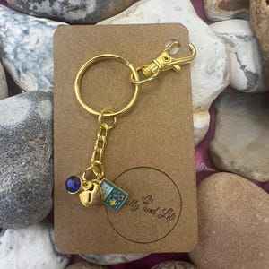 May include: Gold keychain with a blue gem, a gold charm, and a small blue and green charm with a game controller design. The keychain is on a brown background with the text "Polly and Liti" printed on it.